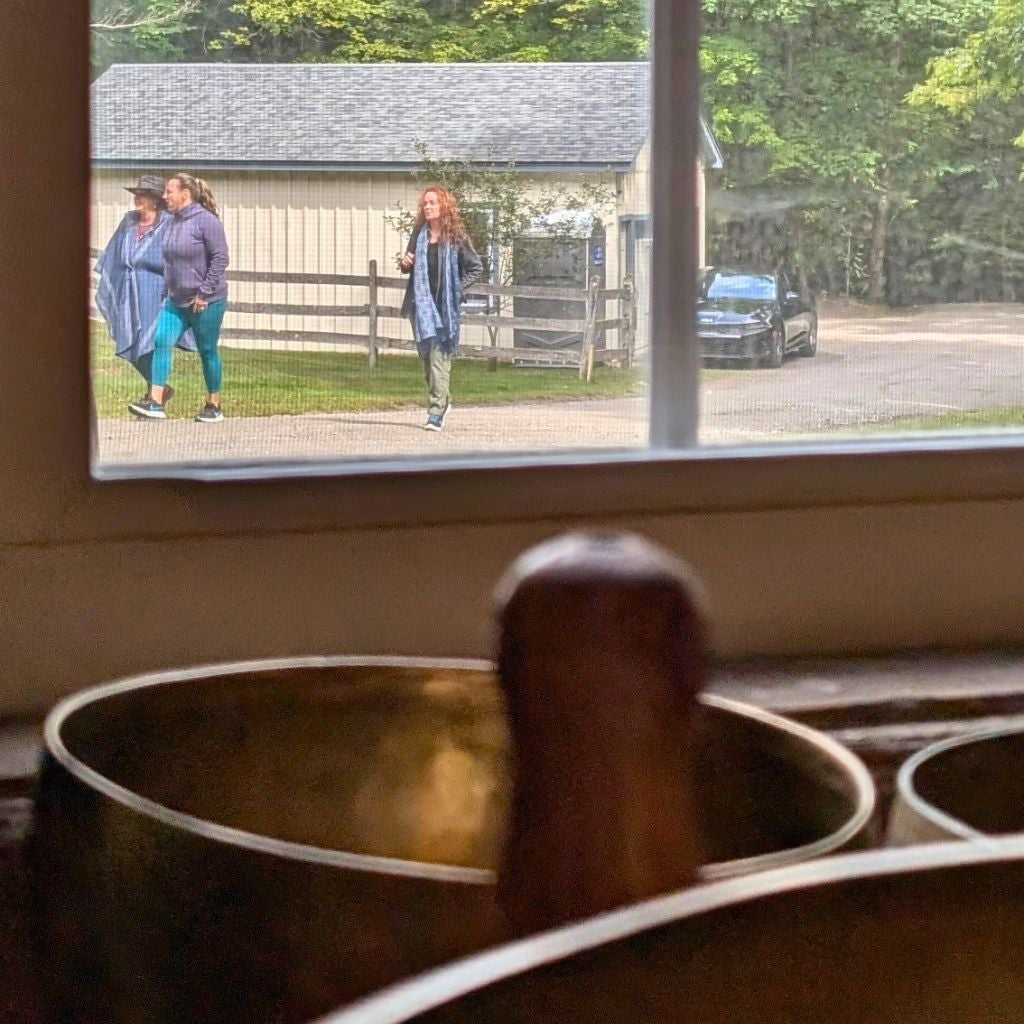 A close-up of metal singing bowls and a wooden mallet in the foreground, with a large window behind them showing two people walking outside near a building and trees.