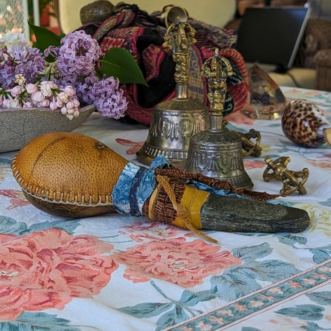 Sound healing instruments including a hide rattle, bells and shell shaker, on a floral-patterned tablecloth. 