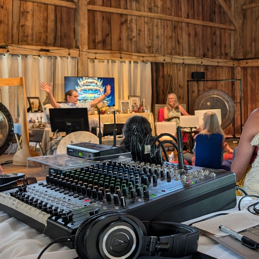Sound mixing console with headphones on a table in a rustic indoor setting with people and equipment in the background.