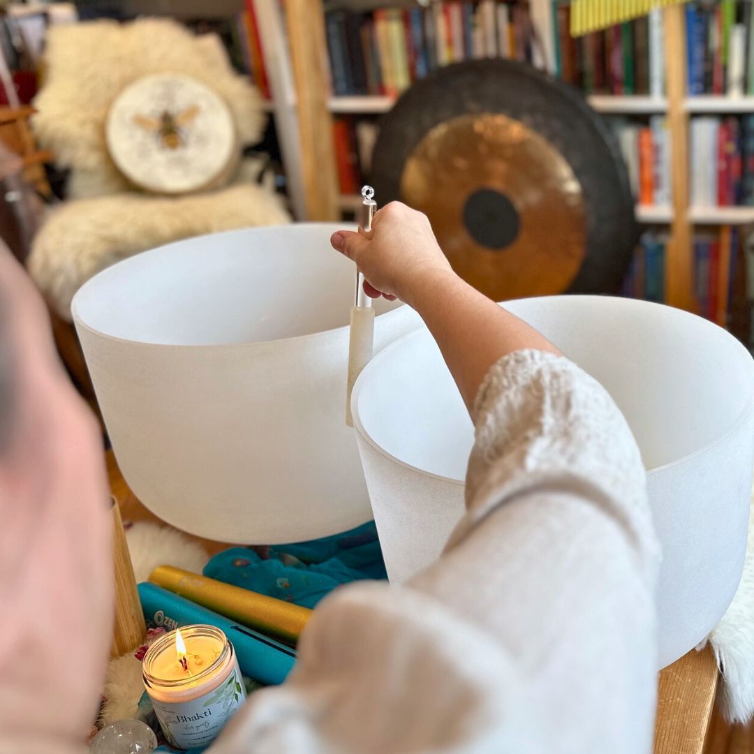 Practitioner playing a crystal singing bowl in a room with a gong and books in the background.
