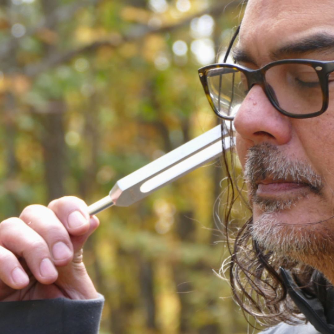 Man holding a tuning fork to his ear outdoors with trees in the background